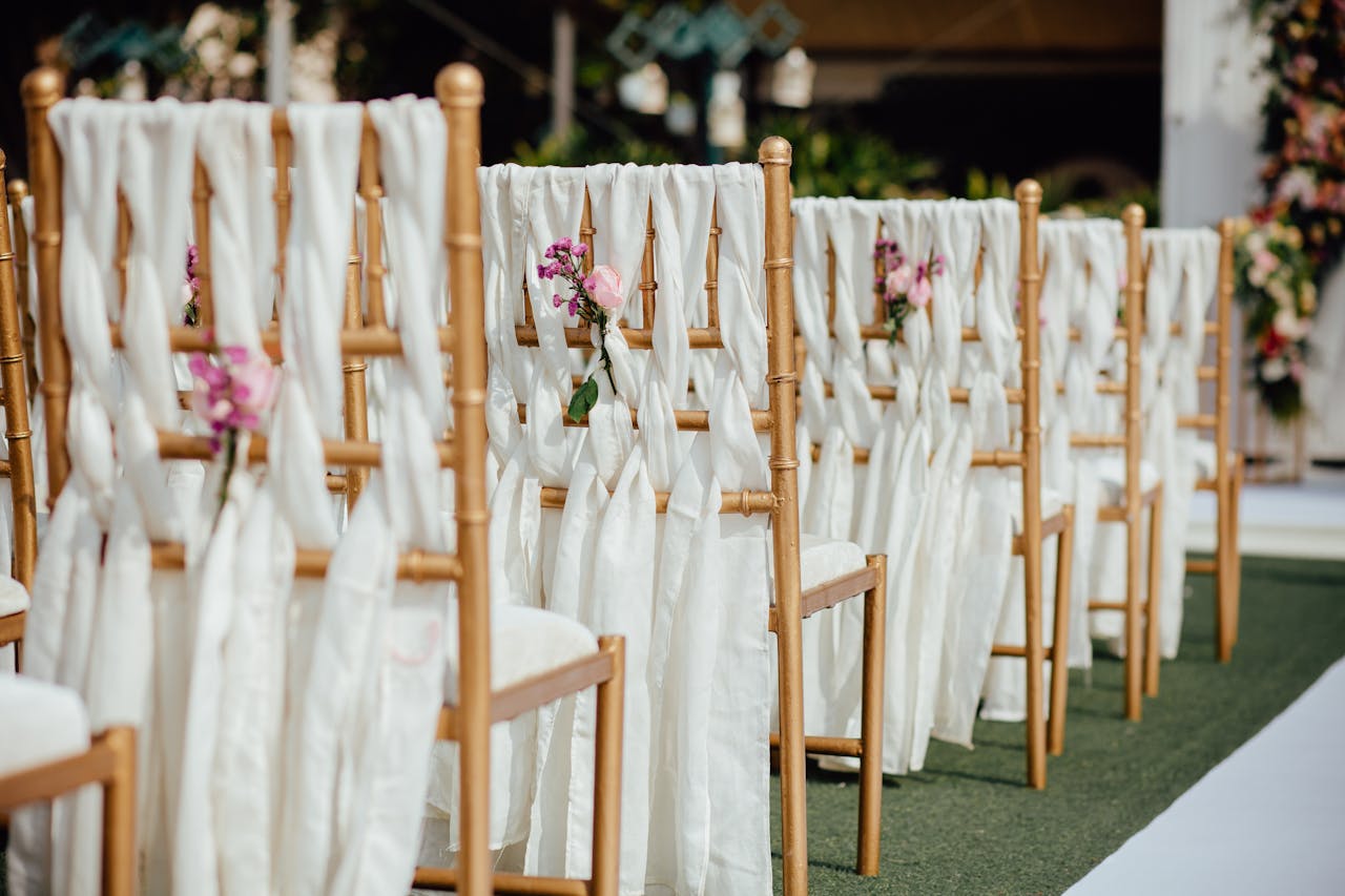 Row of elegant wooden chairs decorated with white fabric and pink flowers for an outdoor wedding destination ceremony.