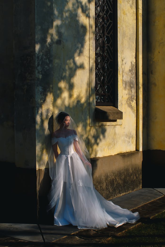 Bride to be in wedding gown poses gracefully against sunlit wall.