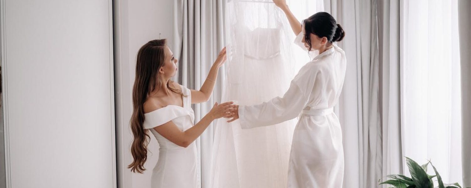 Bride and bridesmaid preparing wedding dress in a bright room.